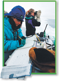 A scientist looks out at the ocean with binoculars, and another records information in a log book.