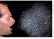 A photograph of a man sneezing. The pathogens released are shown as tiny water particles spraying from the mouth.