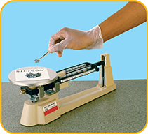 A balance scale with a sample of silicon on the plate and a gloved hand reaching down to pick up silicon with tweezers.
