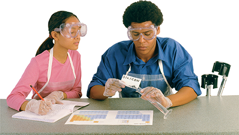 Two students work in a lab. The girl takes notes in a notebook while watching the male student pour silicon into a clear tube.