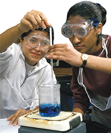 Two students work in a science lab. One student holds a probe steady in a measuring cup filled with liquid while the other student performs the test. 