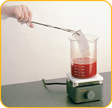 A gloved hand uses tongs to dye a strip of fabric in a measuring glass containing liquid,  kept on a heater.