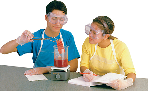 Two students work in a lab. The male student holds a strip of dyed fabric with tongue as it comes out of the hot glass measuring cup. The female student watches and takes notes in a notebook. 