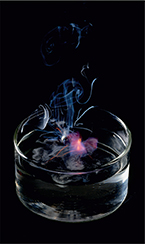 A small glass dish with a spark in the center, releasing smoke against a black background.