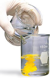 A gloved hand carefully pours liquid from one measuring glass into another. As the liquid is poured in, the liquid in the measuring glass turns yellow.