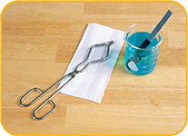 A pair of tongs set on a paper towel next to a solution in a small beaker with a long strip sticking out of the solution.