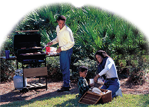 A young family prepares food for a picnic. A man stands at the barbeque grill, while a woman and child get drinks out of a basket. 
