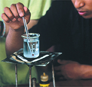 A student dips a thermometer into a glass beaker filled with liquid atop a burner.