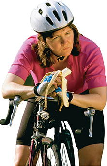 A woman wearing cycling clothes and a helmet, sits on a bicycle, eating a banana.