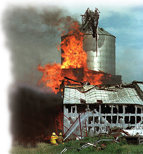 A burning mill covered in flames. Firefighters on the ground try to put it out with a water hose. 