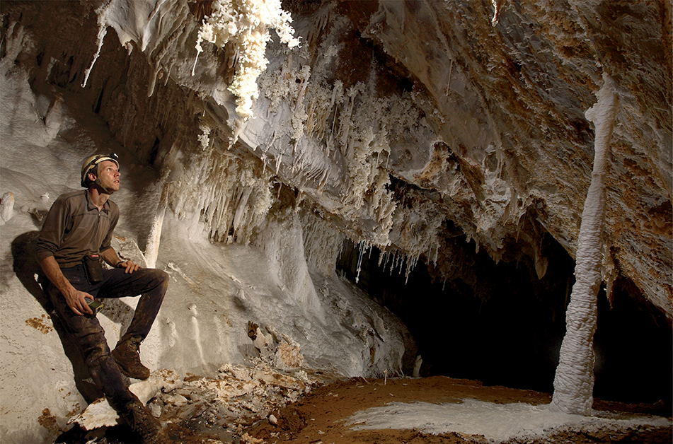 The inside of a cave, with mineral formations growing down from the ceiling and up from the floor. A man wearing a helmet with a torch sits inside.
