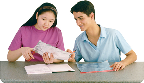 Two students work in a lab. The female student is cutting a piece of tin foil. The male student is watching her. On the table in front of them is a notebook, another sheet of tin foil, and a ruler.