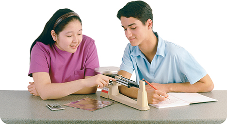 Two students work in a lab. The female student  shows the other student how to use a balance scale. The male student watches her and takes notes in a notebook.