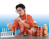 A male student conducts a lab experiment. He drops a liquid into a test tube containing a type of juice. There are three juice bottles next to him on the table, labeled apple juice, Hawaiian pineapple juice, and grapefruit juice.