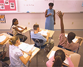 A classroom filled with students working in their workbooks while the teacher lectures at the front of the room. One student is raising his hand.