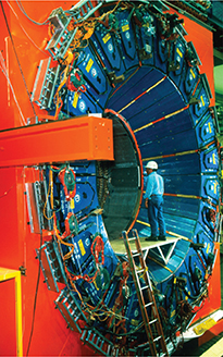 A man stands inside a giant industrial machine that resembles the shape of a tunnel. There is a ladder below the platform where he stands to enter the tunnel-like machine.