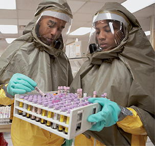 Technicians wearing protective clothing hold a tray of test tubes containing chemicals. 