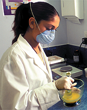 Photo of female wearing a lab coat and mixing solutions in a glass bowl, wearing a face mask.