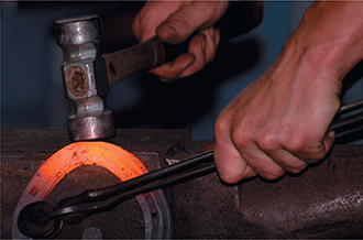 Photo of someone using a hammer to mold and shape a hot piece of metal shaped like a horseshoe.