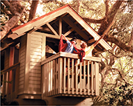 Photo of a wooden treehouse set high in a tree, with two people dressed up in costumes playing on its balcony.