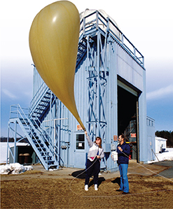 A woman holds a giant weather balloon, as it expands into the air. A man stands next to her holding a box that is connected to the balloon by a cord.