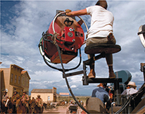 Photo of a man on a lift in the air holding a piece of filming equipment on the set of a movie.