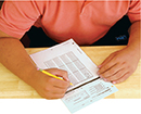 A person holding a pencil, with a sheet of paper in front of them on a desk.  The person is taking a test.