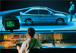 Two engineers conducting an efficiency test on a car. One engineer is outside of the glass enclosed room monitoring the data on machines. The other engineer is inside the glass enclosed room with the car observing how the smoke passes over the car.