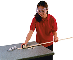A girl wearing safety goggles, leaning over a table with a  meter stick in her hand.  She is using the meter stick to measure a pencil that is taped parallel to and near the edge of the table. There is a spring scale on the table. 