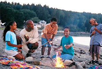 A family of 5 at the bank of a river, toasting marshmallows around a small fire.