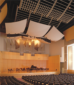 An empty theater inside a music building.  The theater is designed in a way where the audience can hear fully the pitch of different instruments. There are large rectangular tiles hanging from the ceiling, towards the front o the stage.