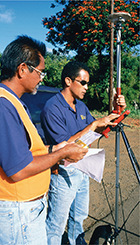 Two surveying technicians using a surveying instrument to collect data.  There are trees in the background.
