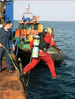 A commercial diver dives into the ocean off the edge of a boat. The diver is dressed in diving gear, with an air tank attached.