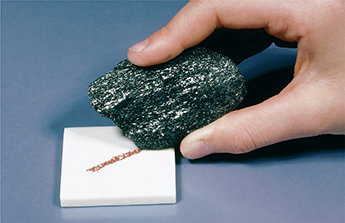 A hand using a piece of rock to make a streak against a small tile.  The streak is a darker color than the tile.