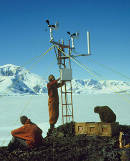 Three scientist setting up a weather station that looks like a large antenna on the frozen tundra of Antarctica.

