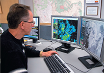 A male meteorologist sitting in front of a computer with a satellite image on the screen.

