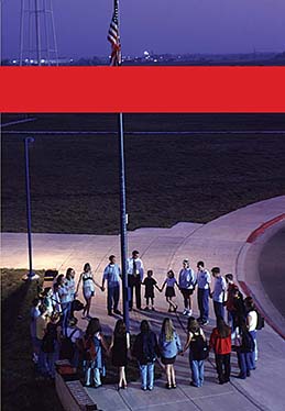 Children stand in a circle around a flagpole with the US flag, with their arms linked.