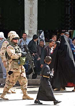 U.S. soldiers with guns walk on the street. Some local men and women watch them, some appear to ignore them.