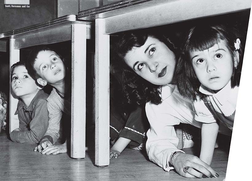 A teacher and some students look out from under school desks.
