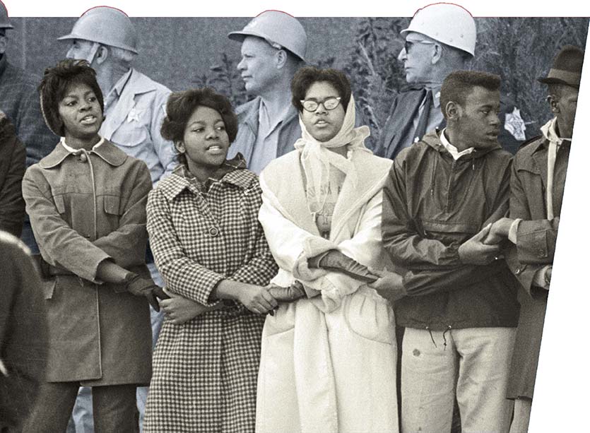 A group of African American protestors holding hands and singing. Behind them are three white men in hardhats.