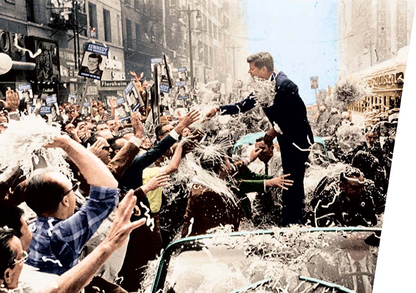 A photograph of John F. Kennedy standing in a convertible automobile, shaking hands with people on the route.