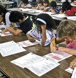 A photo of a class taking a test.
There are test booklets and test answer sheets on desks in front of student.
