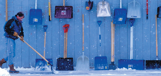 A man shovels snow. Other shovels are displayed on a wall nearby.