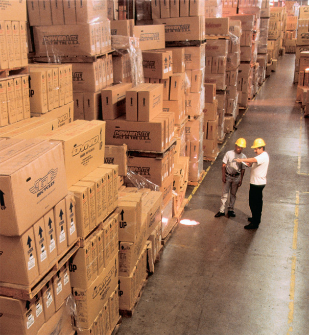 Two men wearing hard hats stand among boxes in a warehouse.