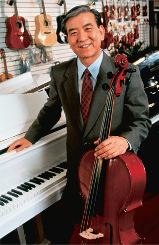 A man holding a cello stands next to a piano, with other musical instruments on the wall behind him