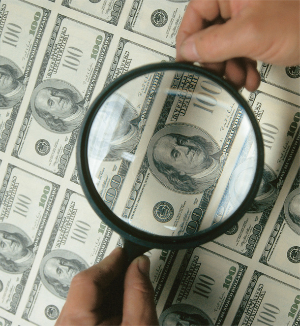 A person holds a magnifying glass to a sheet of 100-dollar bills.
